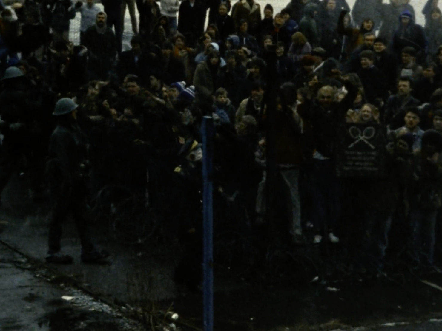 A large crowd of people stand behind a tall metal fence, patrolled by policemen in army helmets.