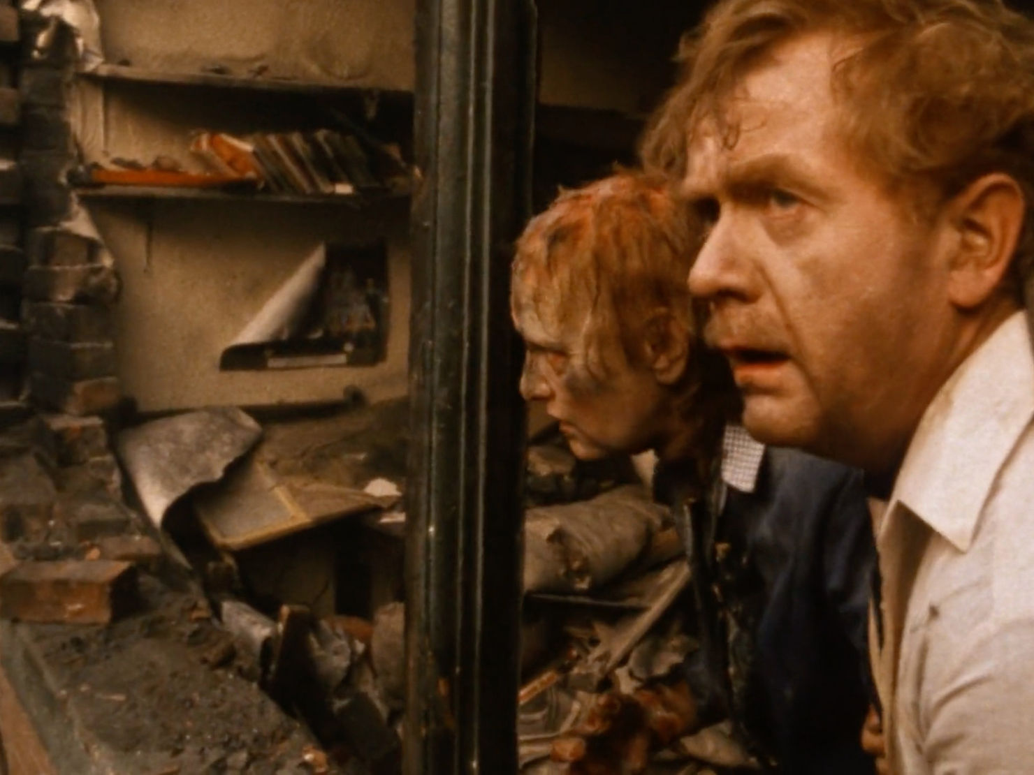 A middle-aged couple, heavily burned, stand in front of their home. Their front wall has been completely demolished and their front room is filled with rubble and ashes.